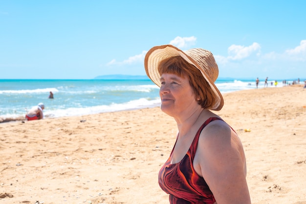 Uma mulher idosa adulta com um chapéu e um maiô na costa arenosa olha para o mar. Viagens e turismo aposentados.