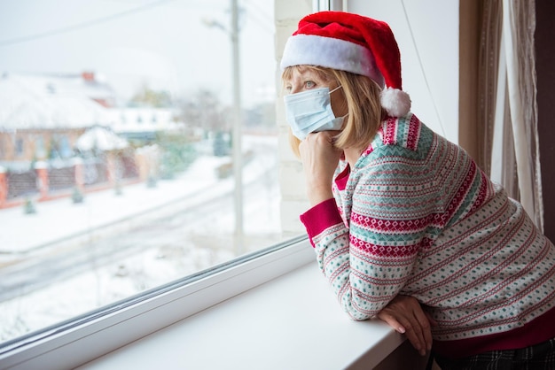 Uma mulher idosa com um chapéu de Papai Noel fica em casa atrás da janela, olhando para fora. Conceito de solidão ou bloqueio devido ao coronavírus covid-19