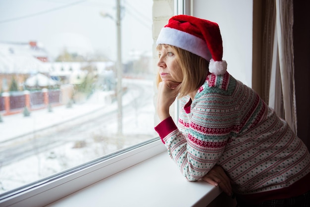 Uma mulher idosa com um chapéu de Papai Noel fica em casa atrás da janela, olhando para fora. Conceito de solidão ou bloqueio