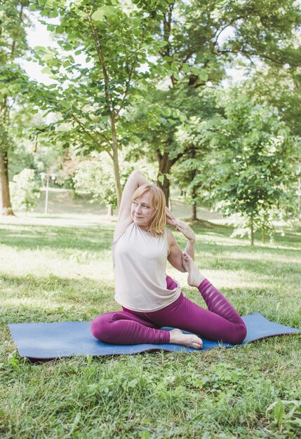 Uma mulher idosa faz ioga no parque em uma esteira. Executa exercícios complexos de Asana e leva uma vida saudável