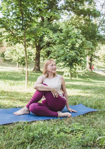 Uma mulher idosa faz ioga no parque em uma esteira. Executa exercícios complexos de Asana e leva uma vida saudável