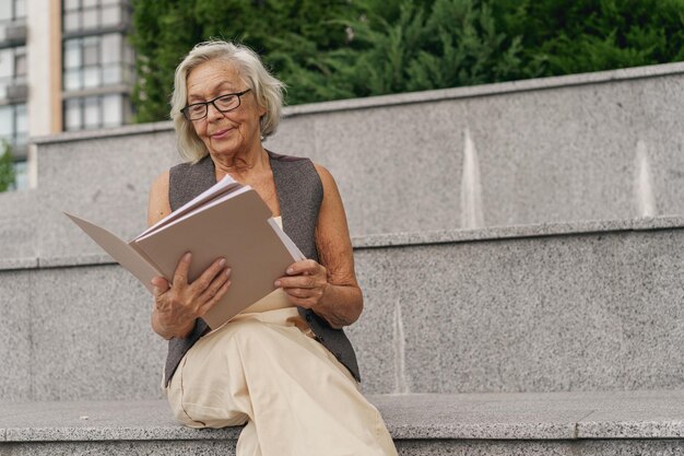 Uma mulher idosa graciosa e elegante está sentada do lado de fora lendo um livro em um cenário urbano