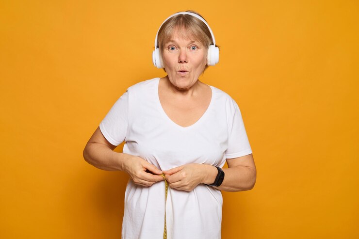 Foto uma mulher idosa parece surpresa enquanto segura uma fita métrica na cintura usando fones de ouvido brancos o fundo amarelo brilhante adiciona um contraste alegre à sua expressão