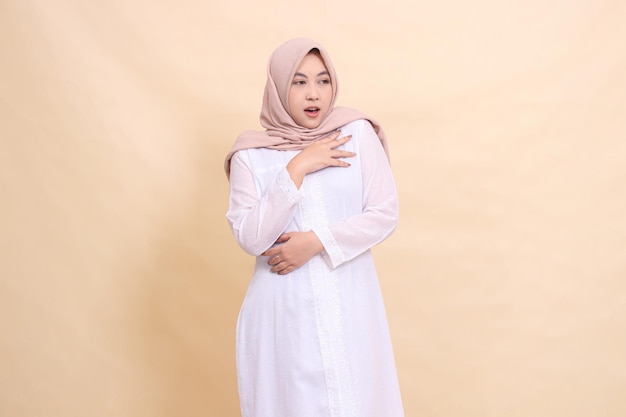Uma mulher indonésia hijab vestindo um vestido branco e um lenço castanho fica franca com as mãos seguradas
