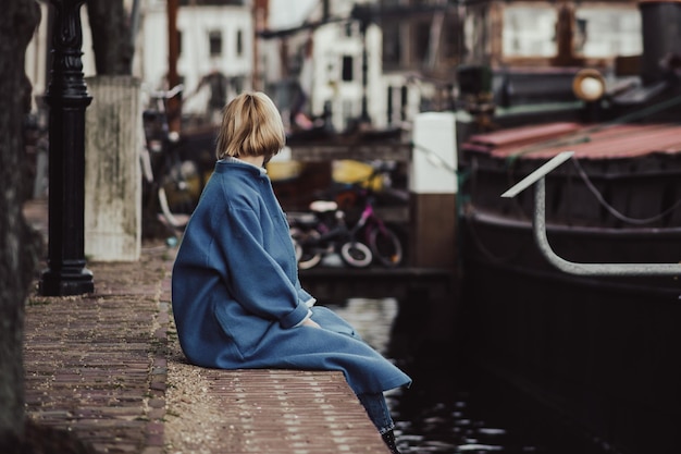 Uma mulher irreconhecível em um elegante casaco azul está sentada na área residencial do cais na Holanda tonificada