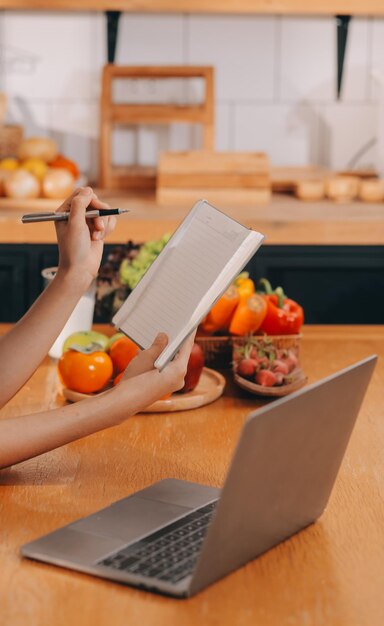 Foto uma mulher jovem com um rosto bonito em uma camisa azul com cabelos longos comendo frutas sentada dentro da cozinha em casa com um laptop e um caderno para relaxar conceito de férias