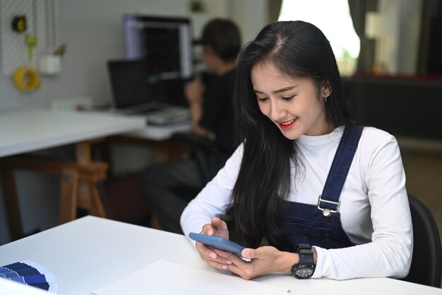 Uma mulher jovem e alegre sentada no escritório e usando o telefone inteligente.