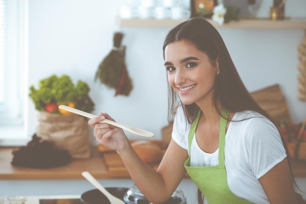 Uma mulher jovem e atraente de cabelos escuros, saboreando uma refeição quente pronta com uma colher de pau em pé e sorrindo perto do fogão da cozinha Conceitos de cozinha e agregado familiar