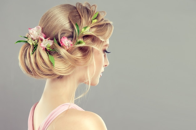 Uma mulher jovem e atraente está demonstrando o cabelo loiro recolhido em um penteado elegante com flores frescas. Arte do cabeleireiro e coloração dos cabelos. Vista de trás.