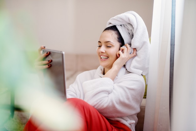 Uma mulher jovem e charmosa está sentada no sofá com um roupão enquanto ouve música pelo fone de ouvido do seu tablet.