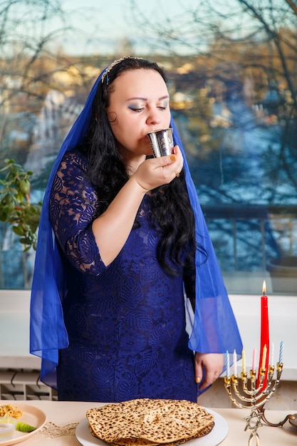 Uma mulher judia com a cabeça coberta por uma capa azul na mesa do Seder da Páscoa bebe vinho do Kidush. Foto vertical