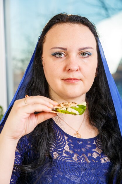 Uma mulher judia com a cabeça coberta por uma capa azul na mesa do seder da páscoa está comendo moror hazeret matzá