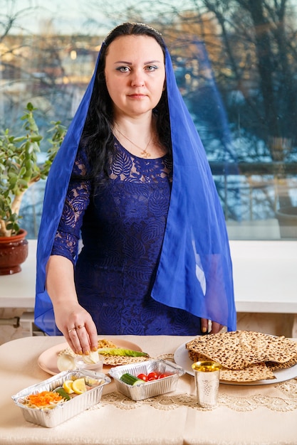 Uma mulher judia com a cabeça coberta por uma capa azul na mesa do Seder da Páscoa executa o mandamento karpas com uma reverência