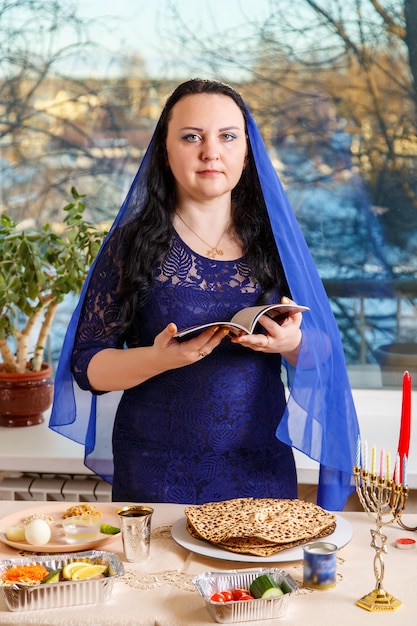 Uma mulher judia com a cabeça coberta por uma capa azul na mesa do seder da páscoa lê a hagadá da páscoa