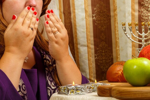 Uma mulher judia com um véu caindo de seu cabelo lê uma bênção nas velas cobrindo o rosto com as mãos na mesa festiva em Rosh Hashaná