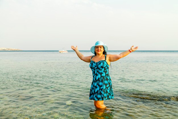 Uma mulher judia feliz em um vestido de verão azul e um chapéu está no mar com água até os joelhos. Foto horizontal