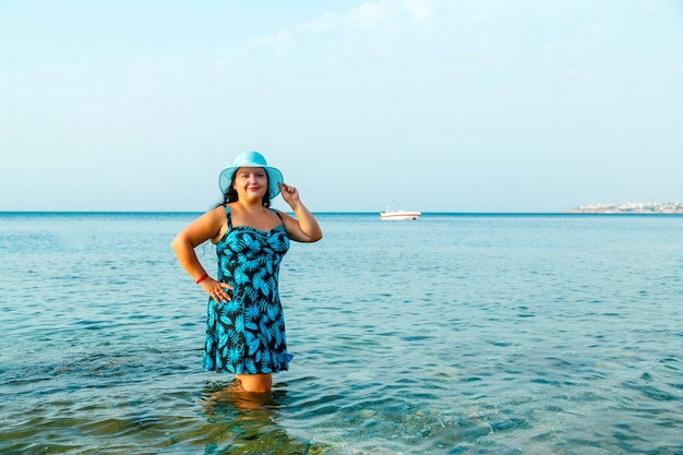 Uma mulher judia feliz em um vestido de verão azul e um chapéu está no mar com água até os joelhos.