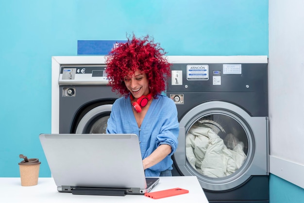 Uma mulher latina com cabelo afro vermelho trabalha com laptop na lavanderia azul