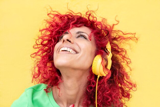 Uma mulher latina feliz com cabelo afro vermelho vestindo uma camiseta verde ouve música com fones de ouvido amarelos olhando sobre um fundo amarelo