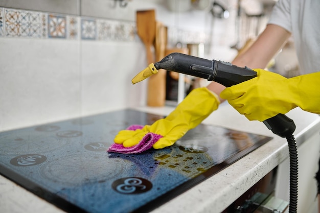 Uma mulher limpa a superfície de um fogão elétrico com um guardanapo depois de processá-lo com um gerador de vapor Limpando a cozinha