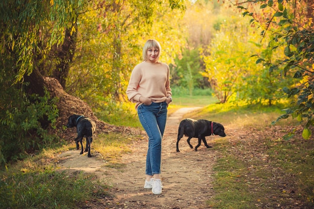 Uma mulher loira com um corte de cabelo curto caminha pela floresta em um suéter de tricô e dois grandes cachorros pretos rottweiler. Caminhada ao ar livre na floresta de outono