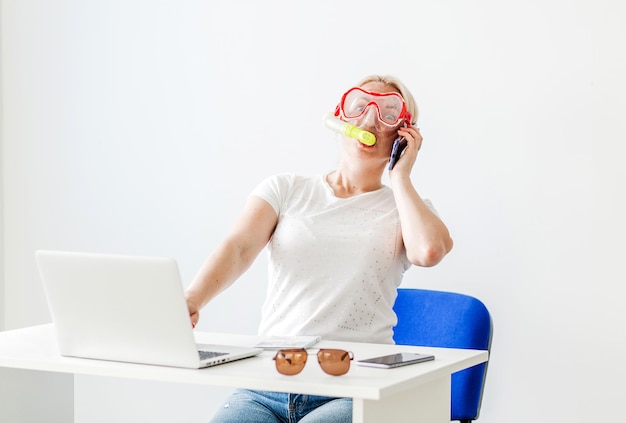 Uma mulher loira com uma máscara de mergulho senta-se no escritório e anuncia alegremente por telefone que vai viajar ou se comunicar com um operador turístico Foto horizontal