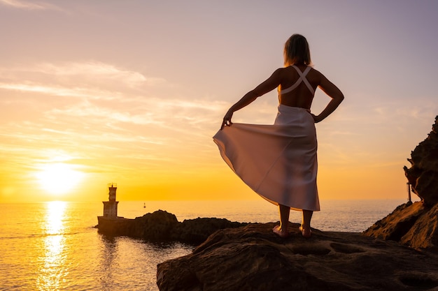 Uma mulher loira de vestido branco ao pôr do sol ao lado de um farol no mar estilo de vida saudável e naturista olhando para o mar