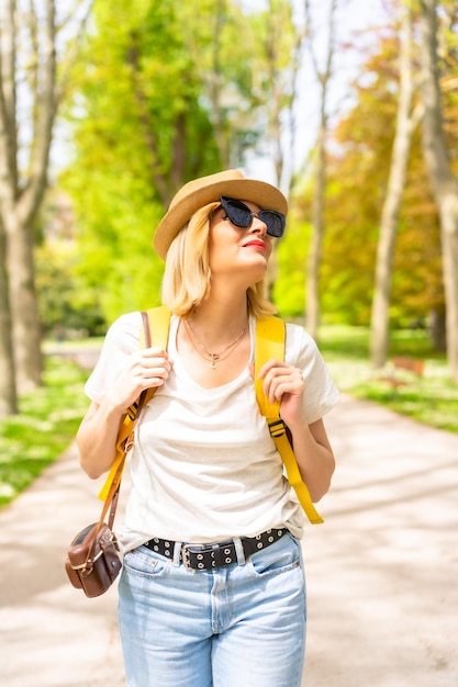 Uma mulher loira turista com um chapéu e óculos de sol andando na primavera em um parque na cidade com a câmera aproveitando o sol