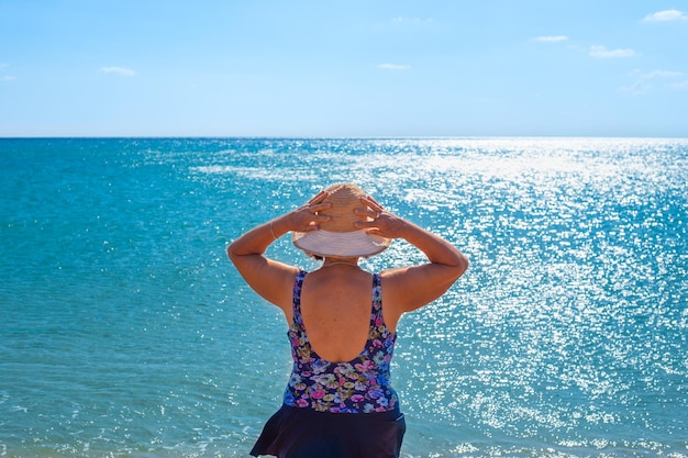 Uma mulher madura de maiô e chapéu olha para o mar azul Viagens e turismo