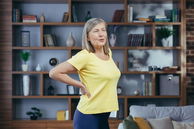 Uma mulher mais velha em casa vestindo uma camiseta amarela está com muita dor