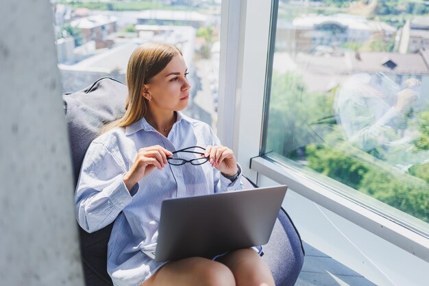 Uma mulher moderna assiste a vídeos em um laptop e bebe café O conceito de distância ou e-learning Uma jovem sorridente se senta em uma cadeira à mesa