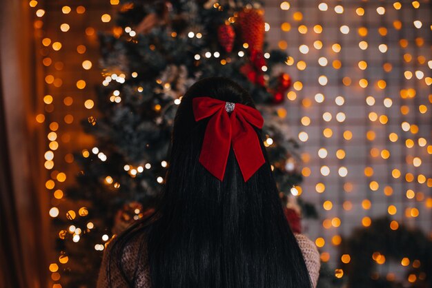 Uma mulher morena com um laço vermelho no cabelo sentada perto da árvore de natal com decorações festivas