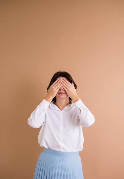 Uma mulher morena de camisa branca fica de pé e cobriu os olhos com as mãos em um fundo bege com um lugar para texto