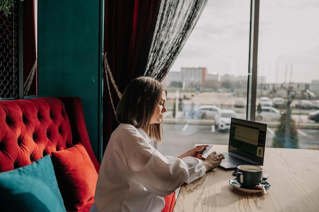 Uma mulher morena de negócios com uma camisa branca está sentada à mesa em um café com um laptop e fazendo compras online com um cartão de plástico