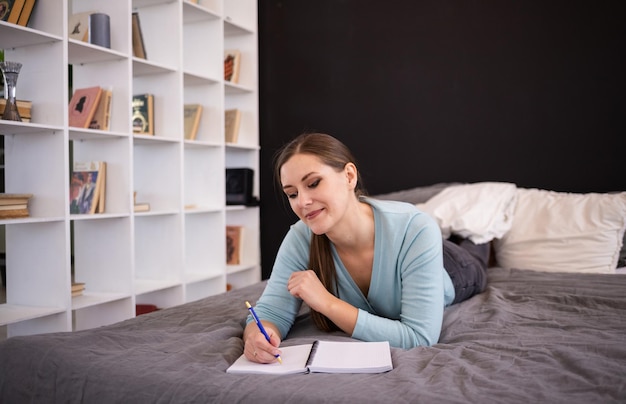 Uma mulher morena entusiasmada deita na cama do quarto e faz anotações em um caderno. mantendo um diário pessoal