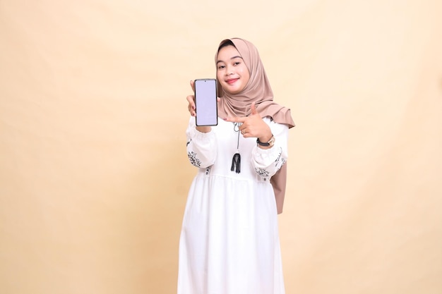 Uma mulher muçulmana asiática adulta usando um hijab sorri mostrando a tela do gadget de seu celular com um polegar