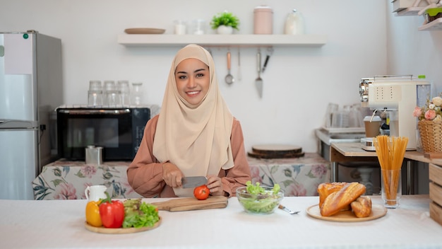 Uma mulher muçulmana asiática sorridente está cozinhando sua mistura de salada saudável em uma mesa na cozinha