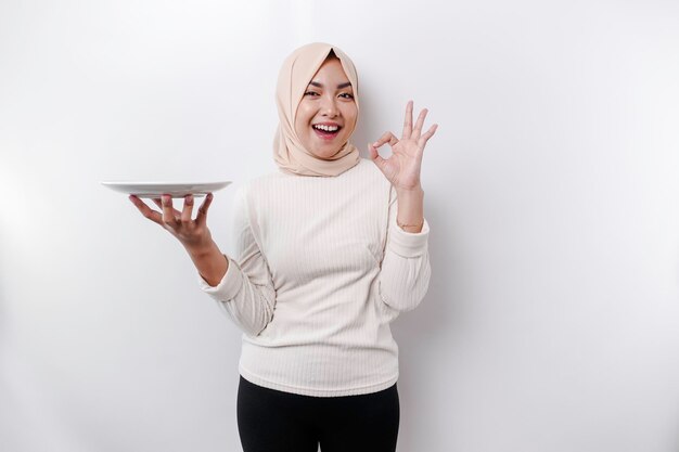 Uma mulher muçulmana asiática sorridente está jejuando e com fome e segurando e apontando para um prato