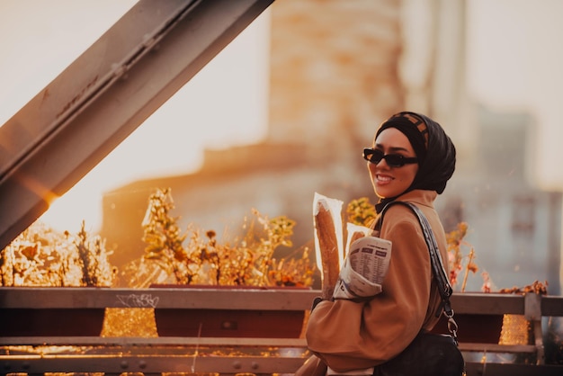 Uma mulher muçulmana moderna com um hijab caminha pela cidade enquanto carrega um jornal e pão na mão.