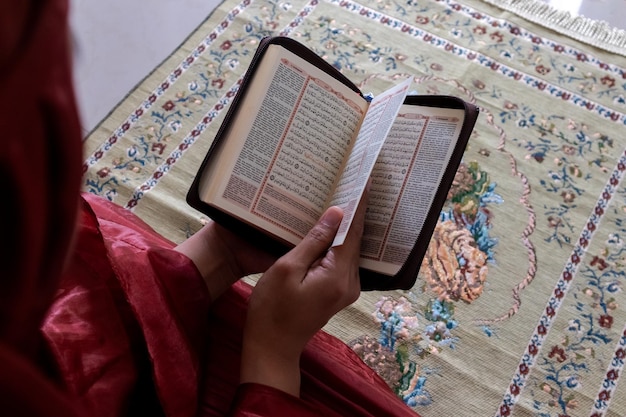Uma mulher muçulmana sentada em um tapete de oração e lendo o Alcorão com tradução indonésia