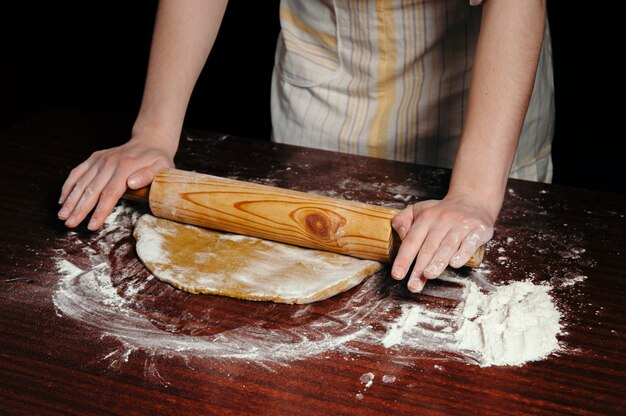 Uma mulher na cozinha está rolando uma massa sobre uma mesa de madeira com um rolo de madeira.