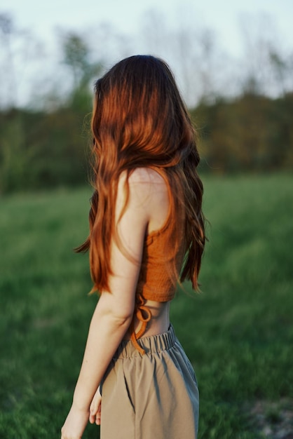 Uma mulher na natureza em um parque verde fica de costas para a câmera e observa o pôr do sol ao ar livre