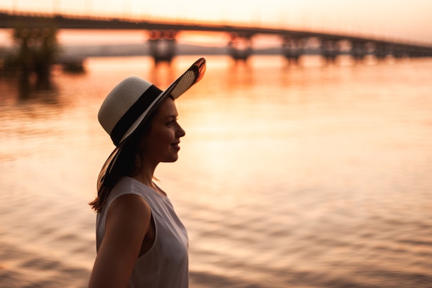 Uma mulher na ponte sobre o rio ao pôr do sol perfil retrato de uma jovem com um chapéu de palha admi ...