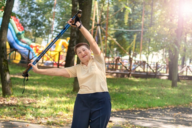 Uma mulher no parque anda nórdica com varas em um dia ensolarado de verão. Mulher sênior de bom humor está envolvida em exercícios matinais.