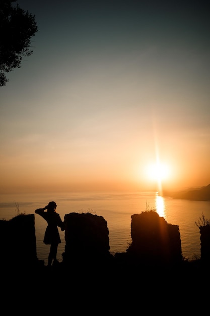 Uma mulher ou um homem de pé sobre uma rocha e olhando diretamente para o pôr do sol à beira-mar O conceito de natureza e beleza Laranja pôr do sol Silhueta ao pôr do sol