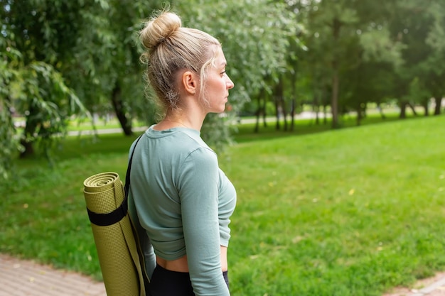 Uma mulher parada no verão no parque com um tapete de ginástica verde para esportes