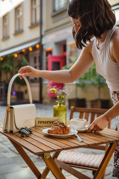 Uma mulher põe a bolsa na mesa e senta-se para comer um croissant e tomar um café