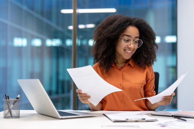 Uma mulher profissional em um escritório sorri enquanto examina papéis em sua mesa com um laptop