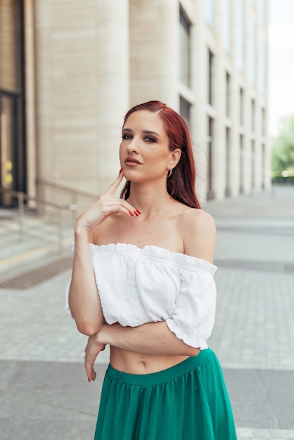 Uma mulher ruiva com roupas de verão olha pensativamente para a câmera na frente de um edifício moderno.