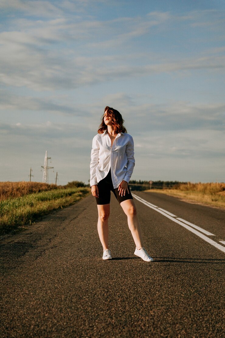 Foto uma mulher ruiva com uma camisa branca caminha ao longo da estrada entre os campos em um dia ensolarado de verão. uma viagem para fora da cidade. foto vertical